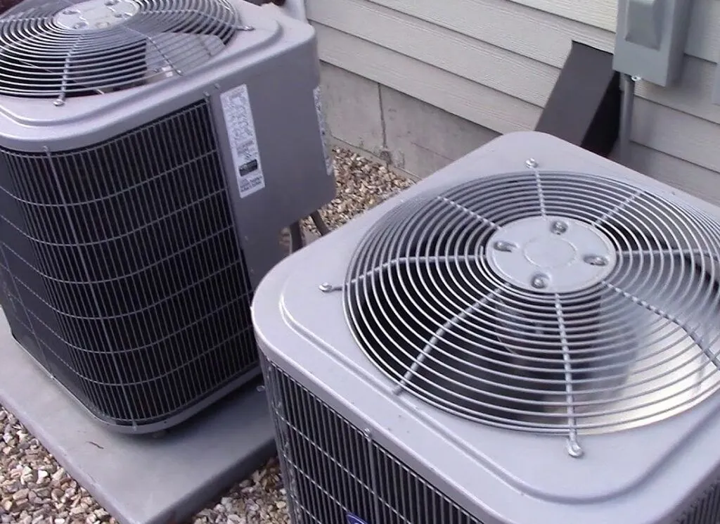 Two residential outdoor air conditioner condenser units side by side on a concrete pad next to a house